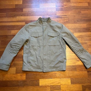 Grön overshirt Massimo Dutti - Säljer en stilren grön jacka från Massimo Dutti. Jackan har en enkel design med två bröstfickor och långa ärmar. Perfekt för en minimalistisk och stilren look.