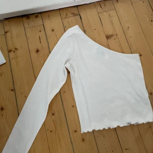 Vit offshoulder topp från Gina Tricot - Säljer en stilren vit offshoulder topp från Gina Tricot. Toppen har en lång ärm och är ribbad med en vågig kant nedtill. 
