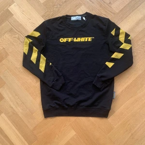 Svart tröja från Off-White med gula detaljer - Säljer en svart tröja från Off-White med gula diagonala ränder på ärmarna och ryggen. Tröjan har Off-White-loggan i gult på magen. Riktigt snygg. XL i barnstorlek så passar S-M