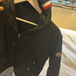 Svart vindjacka från Moncler - Snygg svart vindjacka från Moncler med huva och dragkedja. Oanvänd med tagg. Storlek M