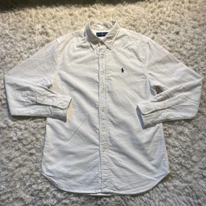 Vit skjorta från Ralph Lauren - Säljer en klassisk vit skjorta från Ralph Lauren med broderad logga i blått på bröstet. Skjortan har långa ärmar och knappar framtill. Perfekt för en stilren look.
