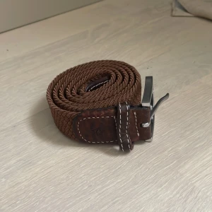 Brunt flätat bälte med silverfärgat spänne - Snyggt brunt flätat bälte med ett stilrent silverfärgat spänne. Perfekt för att ge en extra touch till din outfit. Bältet har vita sömmar som ger en fin kontrast och en klassisk look.