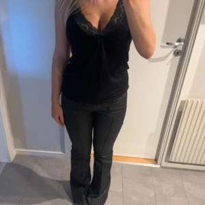 Svart ärmlös topp med spetsdetaljer - Snygg svart ärmlös topp med spetsdetaljer vid halsringningen. Perfekt för en stilren look. Toppen har en v-ringad design som ger en elegant touch.