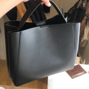 Svart handväska med dragkedja - Snygg svart handväska i skinn med praktisk dragkedja upptill. Perfekt för att hålla dina saker säkra och organiserade. Stilren design som passar till många olika tillfällen.