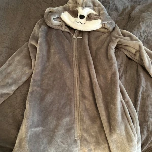 Grå fleece onepiece med djurhuva - Mysig grå fleece onepiece från Mountain Warehouse med en söt djurhuva. Perfekt för kalla dagar hemma. Den har en dragkedja framtill för enkel på- och avtagning.