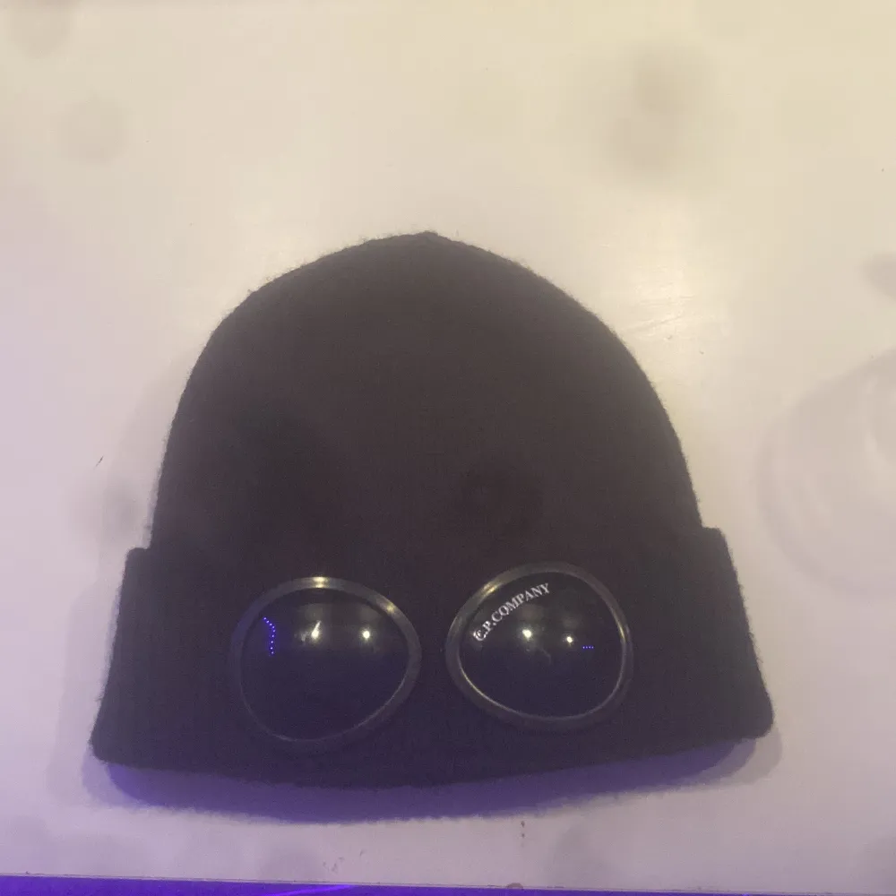 Snygg svart mössa från C.P. Company med unika glasögondetaljer framtill. Perfekt för att ge din outfit en cool touch. Tillverkad i ett mjukt och bekvämt material.. Asusteet.