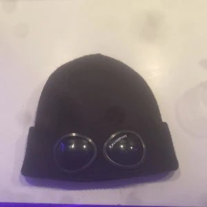 Svart mössa med glasögondetalj från C.P. Company - Snygg svart mössa från C.P. Company med unika glasögondetaljer framtill. Perfekt för att ge din outfit en cool touch. Tillverkad i ett mjukt och bekvämt material.