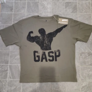 Olivgrön t-shirt från GASP - Säljer en helt oanvänd olivgrön thermal t-shirt från GASP med ett stort tryck av en muskulös siluett och texten 'GASP' på framsidan. Baksidan har texten 'WORK HARD PLAY HARD' i svart. Perfekt för gymmmet. Ursprungligt pris är 900kr