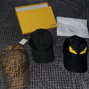 Tre kepsar från Fendi - Säljer tre stiliga kepsar från Fendi. En brun med Fendi-logotypmönster, en svart med gula ögonmotiv och en enkel svart keps. Perfekta för att ge din outfit en lyxig touch. 799kr för en.