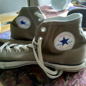 Beige Converse All Star sneakers stl:45 - Säljer ett par klassiska beige Converse All Star sneakers med hög snörning och den ikoniska loggan på sidan. Skorna har en vit sula och är perfekta för en avslappnad stil.