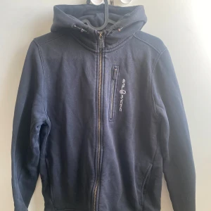 hoodie från Sail Racing - Säljer en hoodie från Sail Racing med dragkedja och huva. Den har en bröstficka med dragkedja och logotyp på bröstet samt på huvan. 