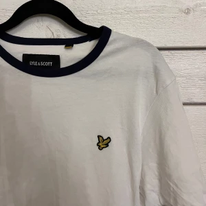 Vit t-shirt från Lyle & Scott - Säljer en stilren vit t-shirt från Lyle & Scott med mörkblå detaljer vid krage och ärmslut. Den har en liten broderad logga på bröstet. Perfekt nu till våren🌸