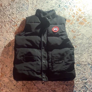  Canada Goose väst - Säljer en svart dunväst från Canada Goose med en klassisk design och varumärkets logga på bröstet. Västen har knappar framtill och två stora fickor. Perfekt för kyliga dagar.