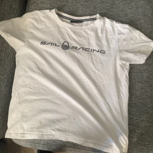 Vit t-shirt från Sail Racing - Vit t-shirt från Sail Racing med svart logotyptryck på framsidan. Klassisk design med rund hals och korta ärmar. Fett tröja inför sommaren 