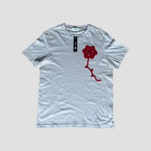 Flower T-Shirt - Snygg vit t-shirt med en unik röd stickad blomma på bröstet. Perfekt för en stilren och modern look. T-shirten har en normal passform och korta ärmar.