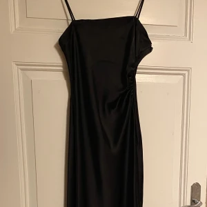 Svart långklänning från Zara - Elegant svart långklänning från Zara med smala axelband. Klänningen har en slät och glansig yta som ger en lyxig känsla. Perfekt för en stilren look.