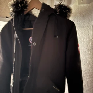 Svart parkajacka med pälsdetaljer - Säljer en stilren svart parkajacka från Canada Goose med en mysig pälsfodrad huva. Jackan har en praktisk dragkedja och knappar framtill. 