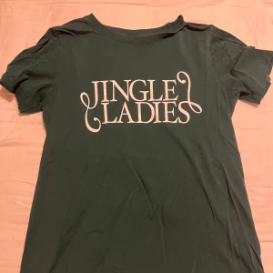 Grön t-shirt från Gina Tricot - Säljer en grön t-shirt från Gina Tricot med texten 'Jingle Ladies' tryckt i vitt på framsidan. Perfekt för att sprida julstämning! T-shirten har en klassisk passform och korta ärmar.