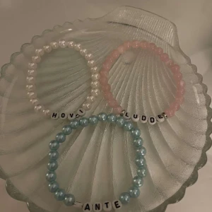 Färgglada pärlarmband med namn - Tre elastiska armband med pärlor i olika färger: vitt, rosa och blått. Varje armband har bokstavspärlor som bildar namn. Perfekt för att ge en personlig touch till din stil.