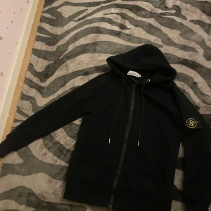 Svart hoodie från Stone Island - Säljer en stilren svart hoodie från Stone Island med dragkedja och huva. Tröjan har en ikonisk logga på ärmen och justerbara snören vid huvan. Perfekt för en casual look. Skriv för mer info