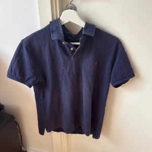 Mörkblå pikétröja från Ralph Lauren - Snygg mörkblå pikétröja från Ralph Lauren med klassisk krage och knappslå. Den har en liten röd broderad logga på bröstet. Perfekt för en stilren look.