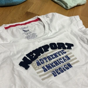 Vit t-shirt från Newport - Säljer en vit t-shirt från Newport med texten 'Authentic American Design' i blått och svart. T-shirten är tillverkad i 100% bomull och har en klassisk rund halsringning. Perfekt för en avslappnad stil.