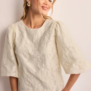 Söt blus från Neo Noir  - Säljer en elegant vit blus med vackert texturerat mönster och trekvartsärmar. Blusen har en fin rosett i ryggen som ger en extra touch av stil. Perfekt för att lyfta vilken outfit som helst!