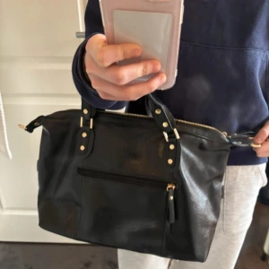 Svart handväska med dragkedja - Helt ny väska, aldrig använd  Säljer då jag tyvärr inte får användning för den  Fina guldiga detaljer och vinröd inuti.            