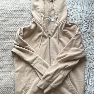 Beige hoodie från H&M - Säljer en beige hoodie från H&M med dragkedja och fickor framtill. Den har en mjuk insida och en justerbar huva med snören. Perfekt för en avslappnad stil. Storlek XS men passar även mig som har S