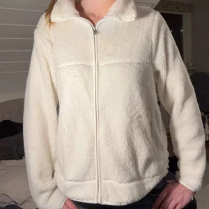 Vit teddyjacka med dragkedja - Mysig vit teddyjacka med dragkedja framtill. Perfekt för kyliga dagar med sin fluffiga och varma design. Jackan har långa ärmar och en hög krage för extra värme. Passar s och m skulle jag säga!