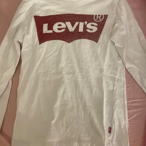 Vit långärmad tröja från Levi's - Säljer en vit långärmad tröja från Levi's med det klassiska röda logotyptrycket på framsidan. Sitter skit snyggt på, de röda trycket är rätt så urtvättad så det har en dämpad röd färg. Annars i bra skick!