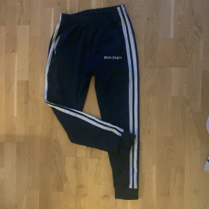 Navy blå byxor från Palm Angels - Snygga mörk blåa byxor från Palm Angels med vita ränder längs sidorna. Byxorna har en elastisk midja och dragsko för justerbar passform. Perfekta för en sportig look.