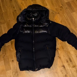 Svart pufferjacka från Polo Ralph Lauren - Säljer en svart pufferjacka från Polo Ralph Lauren. Jackan har en glansig finish med dragkedja framtill och en hög krage. Perfekt för kyliga dagar med sin vadderade design. Den har långa ärmar och praktiska fickor med  dragkedjor. Ny pris 6000-7500kr  Det ända problemet med Jackan är att man inte kan stänga höger fickan. Passsar 185-187. Vid snabb affär kan pris sänkas