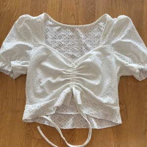 Vit blus med spetsdetaljer -  Supersöt vit blus med spetsdetaljer, har en v-ringad design med knytning framtill och en lätt volang vid nederkanten. 