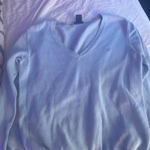 Ljusblå tröja från GANT - Säljer en stilren ljusblå tröja från GANT med v-ringning och ett diskret broderat emblem på bröstet. Perfekt för en avslappnad men ändå snygg look. Tröjan har långa ärmar och är gjord i ett mjukt material.