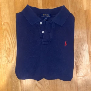 Mörkblå pikétröja från Ralph Lauren - Säljer en klassisk mörkblå pikétröja från Ralph Lauren med korta ärmar och den ikoniska röda broderade loggan på bröstet. Tröjan har en stilren krage och knappar framtill. Perfekt för en avslappnad men stilren look. Den passar dig som är runt 160-170cm. Jag själv är runt 173cm då den är lite för liten för mig. Hör av er om ni har några frågor så svarar jag så snabbt jag kan ! 