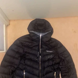 Svart dunjacka från Armani Exchange - Säljer en snygg svart dunjacka från Armani Exchange. Jackan har ett hål på vänster arm men är lagat syns på bilden, annars helt oskadd 