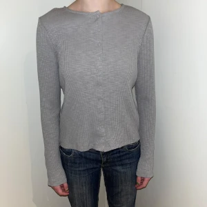 Grå ribbad kofta från Lindex - Säljer en stilren grå ribbad kofta från Lindex. Helt ny, oanvänd! Koftan har långa ärmar och knappar framtill, perfekt för en avslappnad look. Tillverkad i en mjuk bomullsblandning. Passar utmärkt till jeans eller kjol m.m.