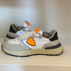 Phillipe model skor - Snygga sneakers från Philippe Model i vitt och beige med en iögonfallande orange detalj på sidan. Skorna har en kombination av mocka och syntetmaterial, samt en robust sula för extra komfort. Perfekta för en stilren look och de är i storlek 43! Nypriset är cirka 2899 kr vilket gör detta till ett riktigt bra pris. Perfekt för sommaren och boxen medkommer:)