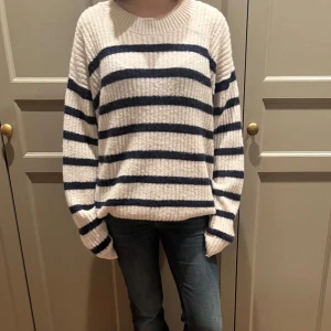 Randig stickad tröja - Mysig stickad tröja med breda marinblå ränder på en krämvit bas. Tröjan har en avslappnad passform med långa ärmar och ribbad struktur. Perfekt för kyliga dagar.