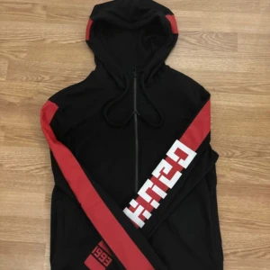 Svart hoodie från Hugo Boss - Snygg svart hoodie från Hugo Boss med röda och vita detaljer. Den har en dragkedja framtill och en stor huva med text. Perfekt för en casual look.