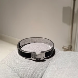Svart armband med ett silver h på - Elegant svart armband  med silverdetaljer och ett h på.  Armbandet kommer i sin originalförpackning med en orange påse och en matchande förvaringspåse. Perfekt för att ge en lyxig touch till din stil. Skriv vid några funderingar och priset är inte spikat