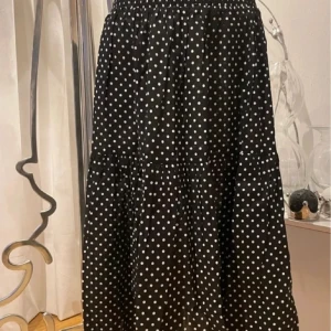 Svart prickig långkjol med smock  - Svart långkjol med vita prickar och bred smock i midjan. Kjolen har volangdetaljer och är luftig med en flowy känsla. Perfekt för dig som gillar en lekfull och trendig stil. Ny med prislappen på . Organic bomull 