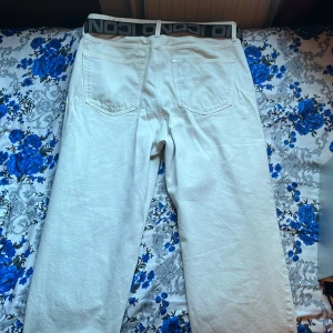 Vita jeans med rak passform - Säljer ett par vita jeans med rak och loose passform. Byxorna har klassiska fem fickor och är tillverkade i bomullstyg. De har en midwaist och kommer med ett svart och grått bälte med grafiskt tryck. Perfekta för dig som gillar clean och enkel stil.