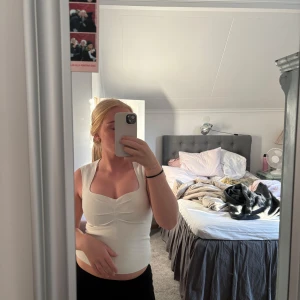 Vit ärmlös linnetopp med snygg urringning - Säljer en vit, ärmlös linnetopp med en cool fyrkantig urringning och lätt rynkade detaljer framtill. Toppen är figurnära och har en clean, stilren look som passar perfekt till jeans eller kjol. Materialet känns mjukt och skönt mot huden.