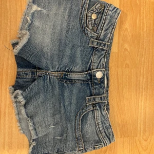Blå jeansshorts från Gina Tricot Young  - Snygga blå jeansshorts från Gina Tricot’s Young kollektion med fransiga kanter och coola fickdetaljer med knappar både fram och bak. femficksmodell, slitna detaljer och grova sömmar ger en avslappnad vibe. Perfekta för varma dagar och chill häng. Säljer då de tyvärr blivit för små för mig!