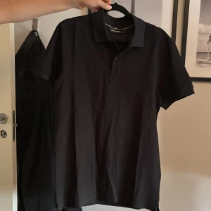 Svart piké från J.Lindeberg - Svart pikétröja från J.Lindeberg med klassisk krage och knappslå. T-shirten är kortärmad och har en stilren, enkel design utan mönster. Materialet är mjuk bomull som känns skönt mot huden. Perfekt för dig som gillar cleana och snygga plagg. Helt ny och oanvänd, endast testad!