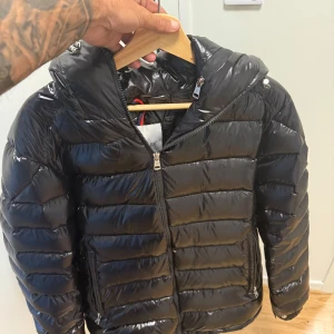 Svart Moncler dunjacka med huva - Säljer en svart, glansig dunjacka från Moncler med huva och dragkedja framtill. Jackan har klassisk quiltad design, två fickor med dragkedja och Moncler-logga på ärmen. Perfekt för kalla dagar och riktigt snygg streetstil. Säljes pågrund av att den inte va som jag tänkte och passa inte min klädstil.Använd 2 gånger förra året och bara hängt i garderoben sen dess. Storlek 3