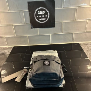 Mörkblå axelväska från Stone Island - helt ny mörkblå axelväska från Stone Island med klassisk kompasslogga på framsidan. Väskan har ett huvudfack med dragkedja, lock med tryckknappar och justerbar axelrem. Tillverkad i slitstark polyester som passar perfekt för att bära dina essentials.