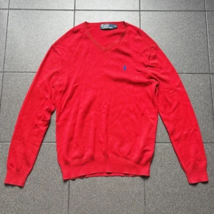 Röd v-ringad tröja från Polo Ralph Lauren - Storlek: S Snygg röd v-ringad tröja från Polo Ralph Lauren med blå broderad logga på bröstet. Tröjan är stickad i mjuk bomull och har långa ärmar samt ribbade muddar vid hals, ärmslut och nederkant. Perfekt för dig som gillar klassisk stil med en färgklick.